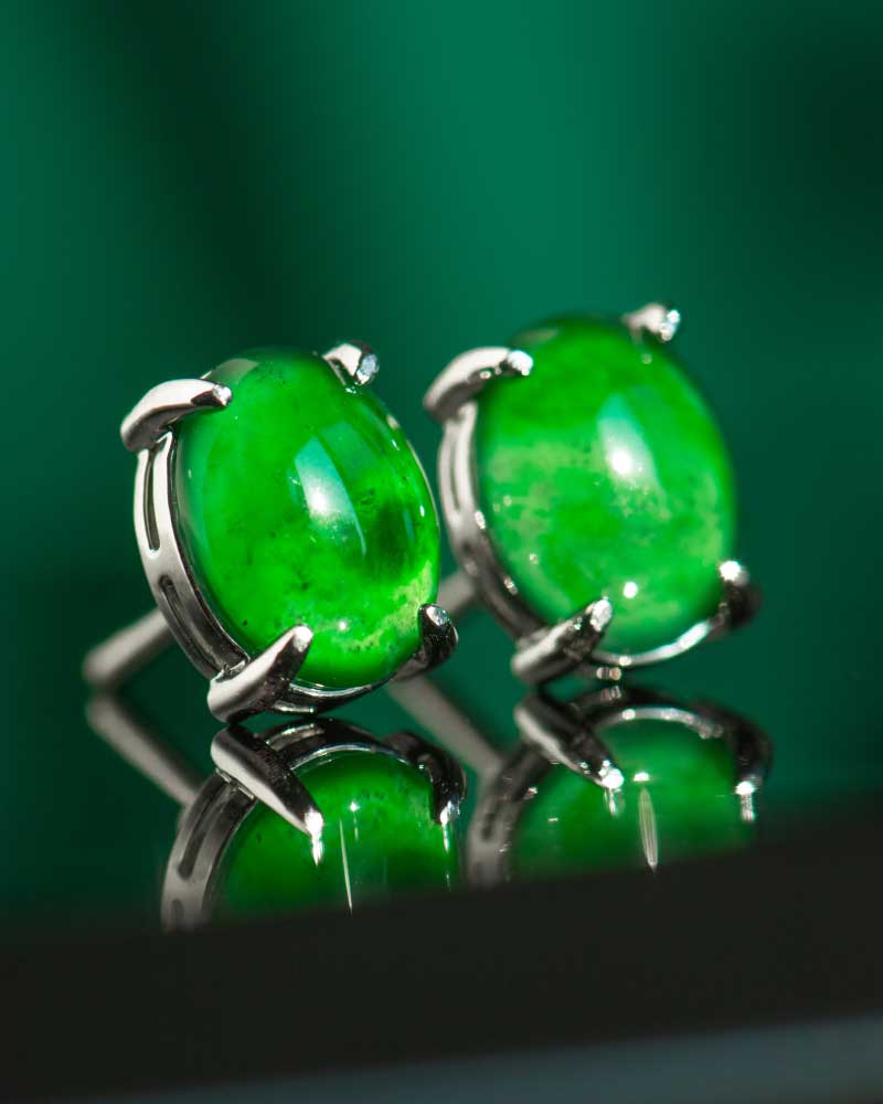 A pair of Vivid Green Jadeite Stud Earrings with round cabochon gemstones set in 925 sterling silver, displayed on a green background with visible reflections.