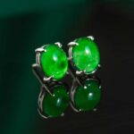 The Vivid Green Jadeite Stud Earrings feature round cabochon gems in 925 sterling silver, showcased on a glossy black surface with reflections beneath them.