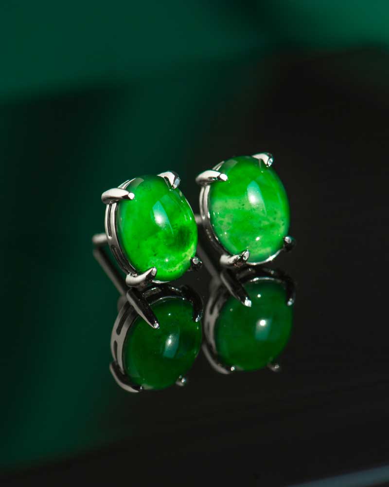 The Vivid Green Jadeite Stud Earrings feature round cabochon gems in 925 sterling silver, showcased on a glossy black surface with reflections beneath them.