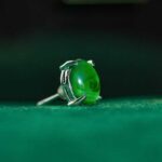 A close-up of the Vivid Green Jadeite Stud Earrings featuring round cabochon gemstones set in 925 sterling silver, displayed on a soft green surface with a dark green blurred background.