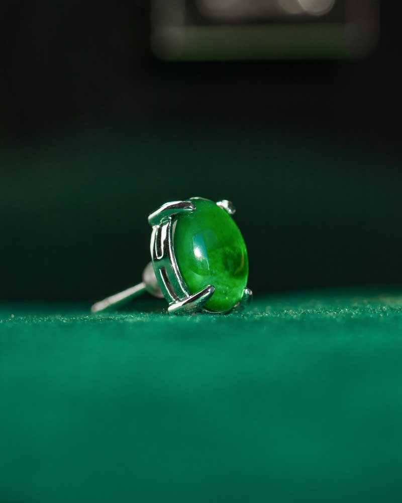 A close-up of the Vivid Green Jadeite Stud Earrings featuring round cabochon gemstones set in 925 sterling silver, displayed on a soft green surface with a dark green blurred background.