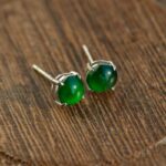 Vivid Green Jadeite Stud Earrings feature round cabochon beads set in silver, showcased on a textured wooden surface.