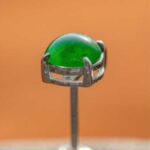 A close-up of Vivid Green Jadeite Stud Earrings featuring round cabochon beads in a silver setting, with a blurred orange background.