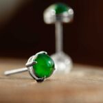 A close-up of Vivid Green Jadeite Stud Earrings—round cabochon beads set in silver—with one earring lying on its side in the foreground and the other upright in the blurred background.