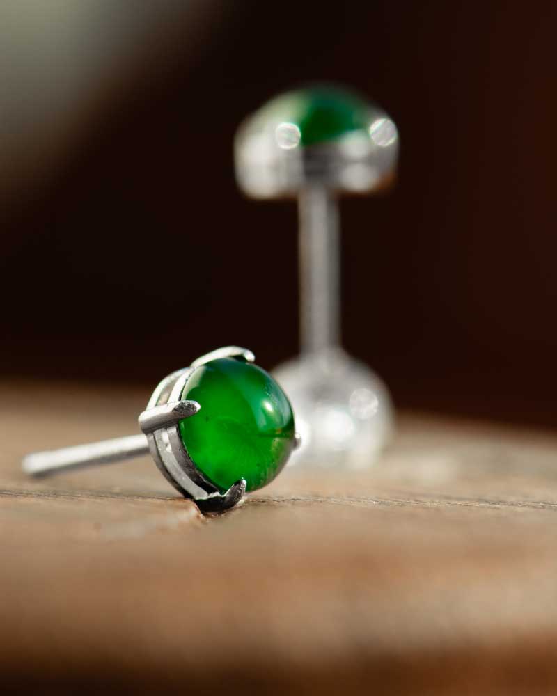 LYU_4971 A close-up of Vivid Green Jadeite Stud Earrings—round cabochon beads set in silver—with one earring lying on its side in the foreground and the other upright in the blurred background.