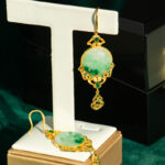 The Jadeite Drop Earring features green circular jade stones in Chinese-inspired gold-plated filigree settings, displayed on a white stand with one earring hanging and the other flat against a dark green background.
