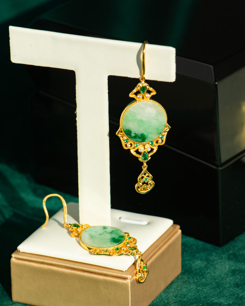 The Jadeite Drop Earring features green circular jade stones in Chinese-inspired gold-plated filigree settings, displayed on a white stand with one earring hanging and the other flat against a dark green background.