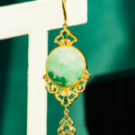 The Jadeite Drop Earring features Chinese-inspired gold-plated 925 sterling silver filigree with emerald-green gemstones and a large round jade centerpiece, displayed against a teal background.