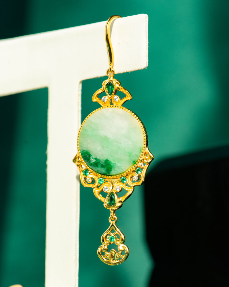 The Jadeite Drop Earring features Chinese-inspired gold-plated 925 sterling silver filigree with emerald-green gemstones and a large round jade centerpiece, displayed against a teal background.