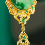 Close-up of the Jadeite Drop Earring—Chinese-inspired filigree in gold-plated 925 sterling silver, with a large green jade stone, accented by small green and white gemstones and a decorative dangling gold element.