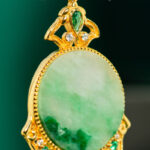 A close-up of the Jadeite Drop Earring, a Chinese-inspired filigree piece in gold-plated 925 sterling silver, featuring an ornate design with an oval green jadeite stone accented by smaller clear and green stones.