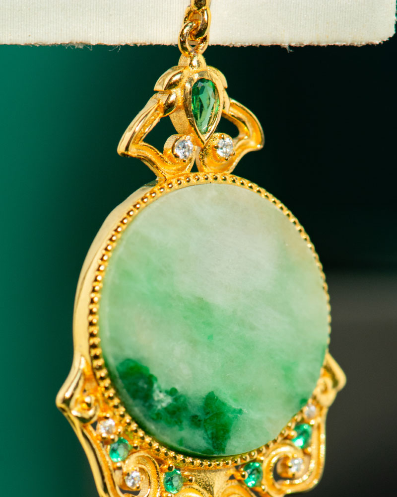 A close-up of the Jadeite Drop Earring, a Chinese-inspired filigree piece in gold-plated 925 sterling silver, featuring an ornate design with an oval green jadeite stone accented by smaller clear and green stones.