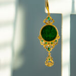 The Deep Green Jadeite Drop Earring features gold-plated 925 silver with Chinese-inspired filigree, a large round jadeite center, small green gems, and hangs against a softly lit backdrop.