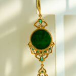 The Deep Green Jadeite Drop Earring features Chinese-inspired filigree in gold-plated 925 sterling silver, with a large round green jadeite center, smaller gemstones, and a dangling design against a light background.