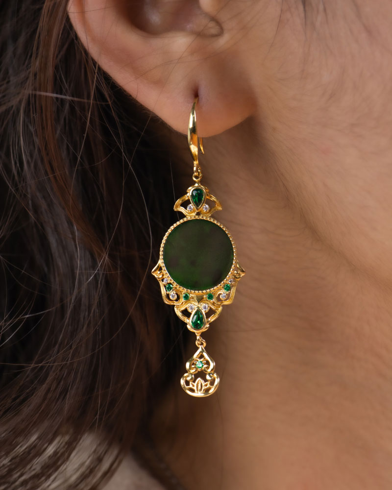 LYU_5071 A close-up of a person’s ear wearing the Deep Green Jadeite Drop Earring—Chinese-inspired filigree in gold-plated 925 sterling silver, with a circular jade centerpiece and teardrop accents.