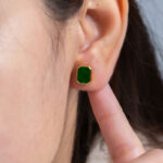 A close-up of a person’s ear wearing a Vivid Green Jadeite Stud Earring with a rectangular flat inlay, set in gold-plated 925 sterling silver. They touch their earlobe with their index finger; the background is plain and blurred.