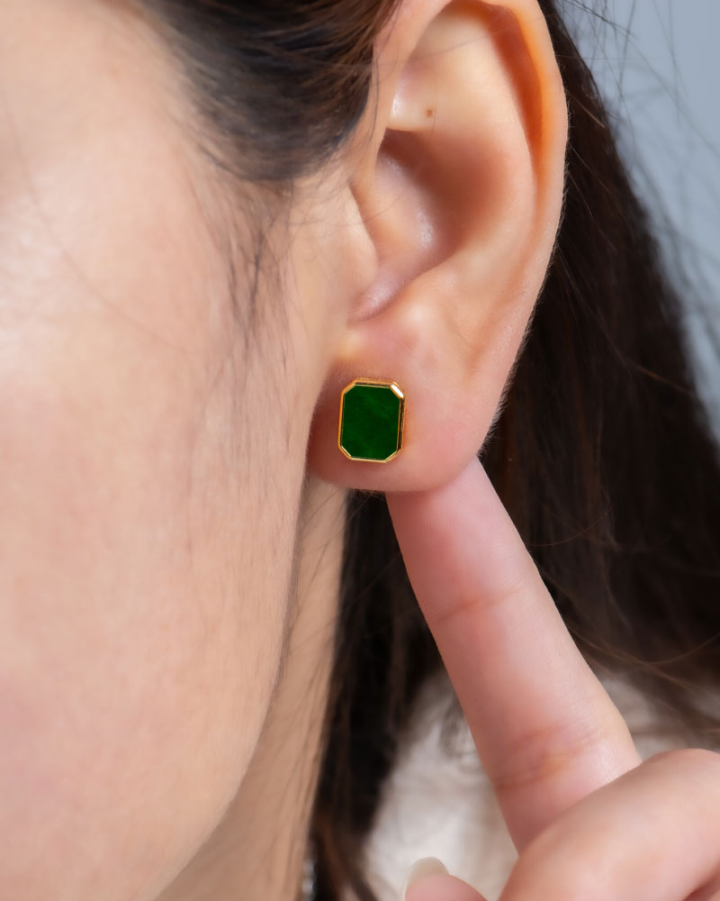 A close-up of a person’s ear wearing a Vivid Green Jadeite Stud Earring with a rectangular flat inlay, set in gold-plated 925 sterling silver. They touch their earlobe with their index finger; the background is plain and blurred.