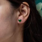 A close-up of a woman's ear wearing Vivid Green Jadeite Stud Earrings—heart-cut with custom faceting, set in 925 sterling silver. Her dark brown hair is partially visible.