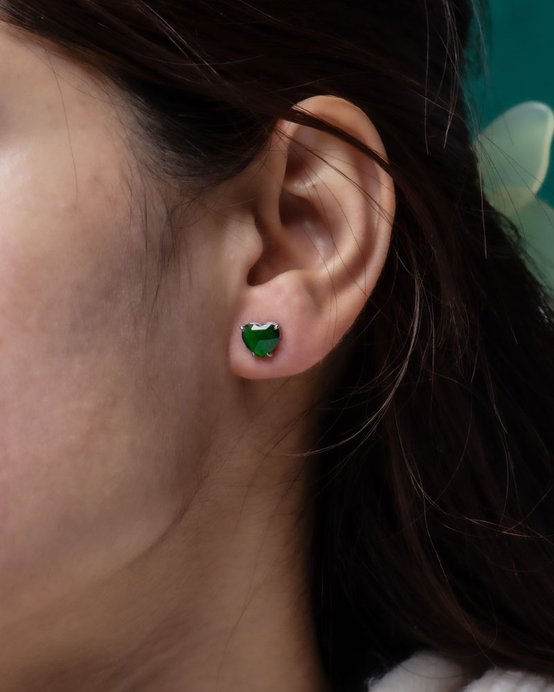 A close-up of a woman's ear wearing Vivid Green Jadeite Stud Earrings—heart-cut with custom faceting, set in 925 sterling silver. Her dark brown hair is partially visible.