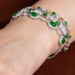 A close-up of a wrist with two silver bracelets, including the Green & Greenish White Jadeite Halo Station Bracelet—oval cabochons set in polished sterling silver—against a brown background.