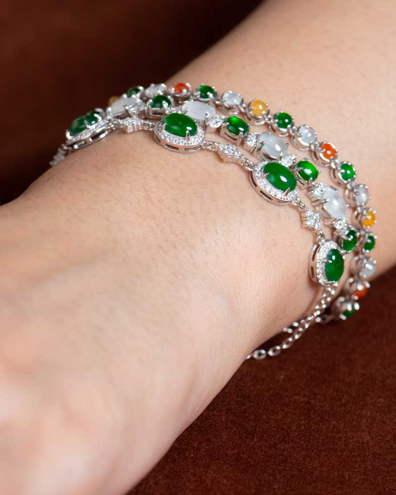 LYU_5155 A close-up of a wrist with two silver bracelets, including the Green & Greenish White Jadeite Halo Station Bracelet—oval cabochons set in polished sterling silver—against a brown background.
