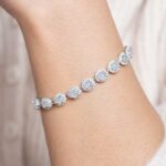 A close-up of a wrist wearing the Greenish White Jadeite Halo Tennis Bracelet, featuring round jadeite gems encircled by clear crystals. The background is cream-colored textured fabric.