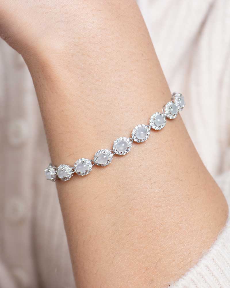 LYU_5164 A close-up of a wrist wearing the Greenish White Jadeite Halo Tennis Bracelet, featuring round jadeite gems encircled by clear crystals. The background is cream-colored textured fabric.