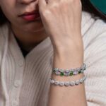 A woman in a cream sweater rests her chin on her hand, showcasing the Greenish White Jadeite Halo Tennis Bracelet with green stones on her wrist and a large gemstone ring on her finger.