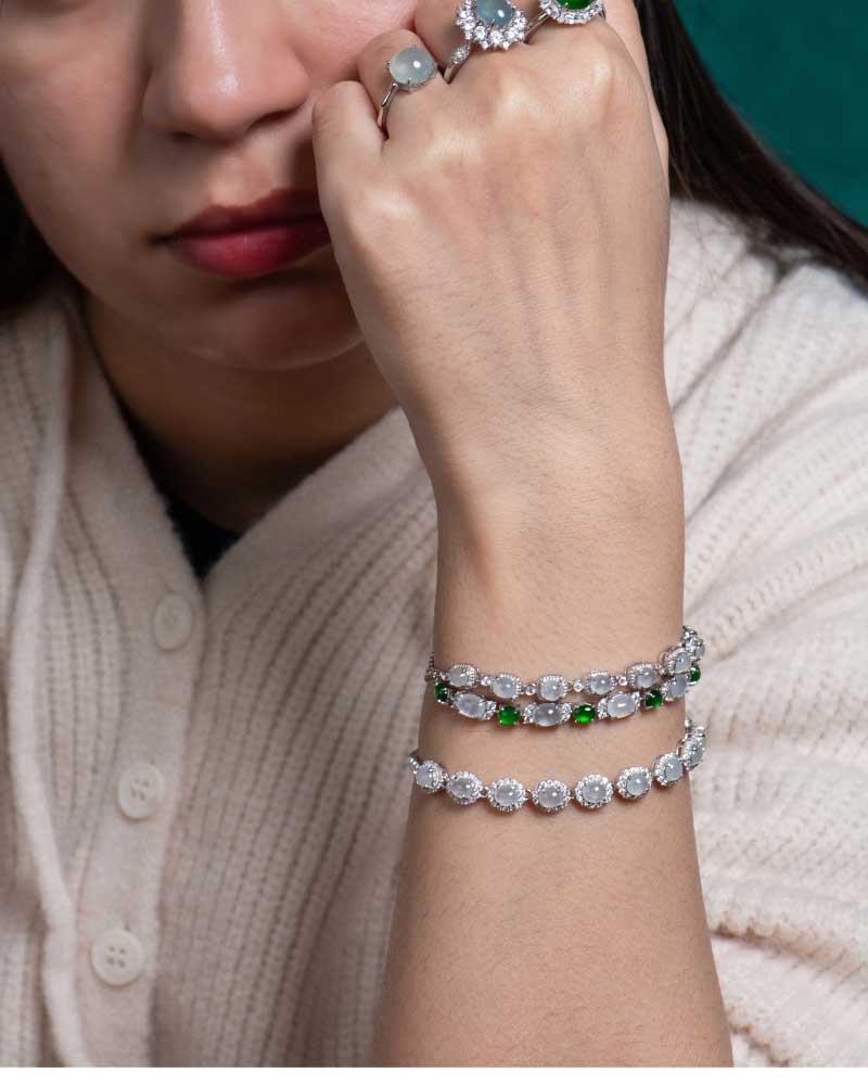 LYU_5168 A woman in a cream sweater rests her chin on her hand, showcasing the Greenish White Jadeite Halo Tennis Bracelet with green stones on her wrist and a large gemstone ring on her finger.