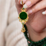A hand with manicured nails holds the Deep Green Jadeite Drop Earring, a gold-plated 925 silver piece with Chinese-inspired filigree and a dark green stone. The person also wears a matching green beaded bracelet.