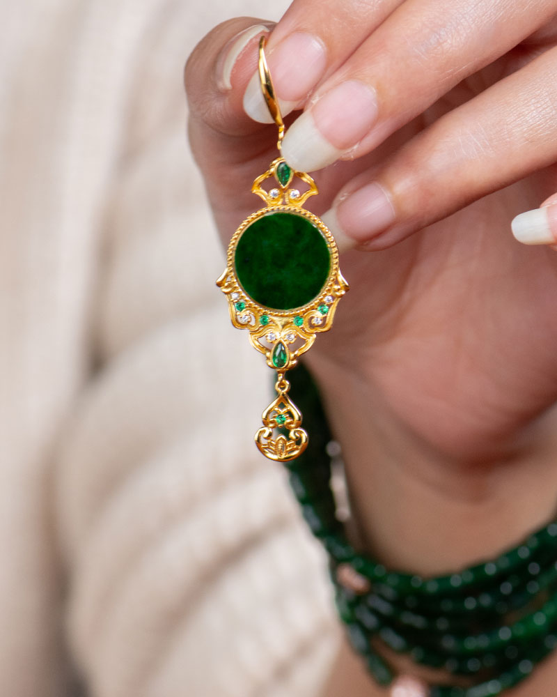 LYU_5184 A hand with manicured nails holds the Deep Green Jadeite Drop Earring, a gold-plated 925 silver piece with Chinese-inspired filigree and a dark green stone. The person also wears a matching green beaded bracelet.