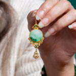 A hand with manicured nails holds a Jadeite Drop Earring—Chinese-inspired filigree in gold-plated 925 sterling silver with a round green jade stone—against a cream sweater backdrop.