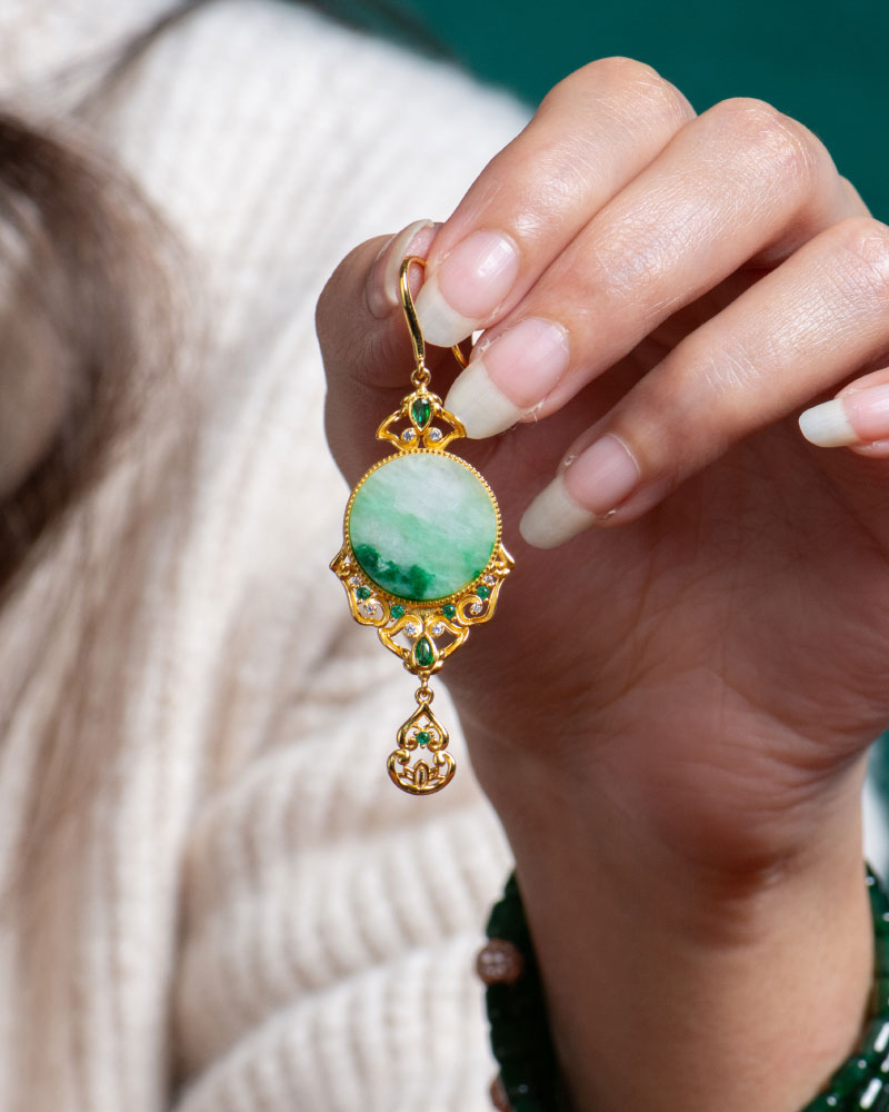 A hand with manicured nails holds a Jadeite Drop Earring—Chinese-inspired filigree in gold-plated 925 sterling silver with a round green jade stone—against a cream sweater backdrop.
