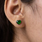 A close-up shows an ear adorned with a sparkling Vivid Green Jadeite Heart-Cut Stud Earring, set in 925 Sterling Silver with custom faceting. Dark brown hair is partially visible in the background.