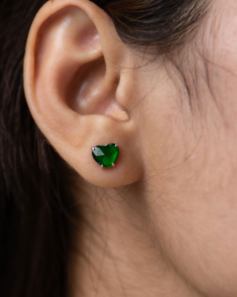 A close-up shows an ear adorned with a sparkling Vivid Green Jadeite Heart-Cut Stud Earring, set in 925 Sterling Silver with custom faceting. Dark brown hair is partially visible in the background.