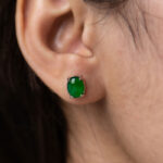 A close-up of a person’s ear wearing a Vivid Green Jadeite Stud Earring featuring a round cabochon gemstone set in 925 sterling silver.