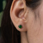 Close-up of a person with straight brown hair tucked behind the ear, wearing Vivid Green Jadeite Stud Earrings – round cabochon beads in a silver setting.