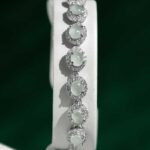 The Greenish White Jadeite Halo Tennis Bracelet showcases oval pale green jadeite gems, each surrounded by sparkling clear crystals, elegantly presented on a white stand with a dark green background.