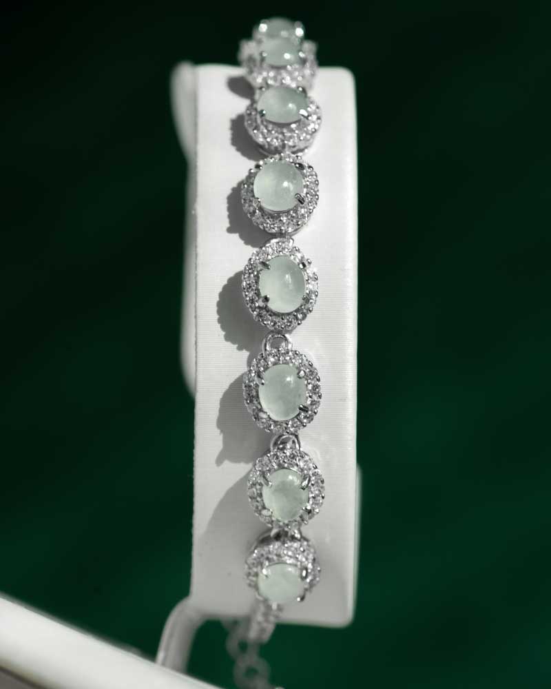 LYU_5277 The Greenish White Jadeite Halo Tennis Bracelet showcases oval pale green jadeite gems, each surrounded by sparkling clear crystals, elegantly presented on a white stand with a dark green background.