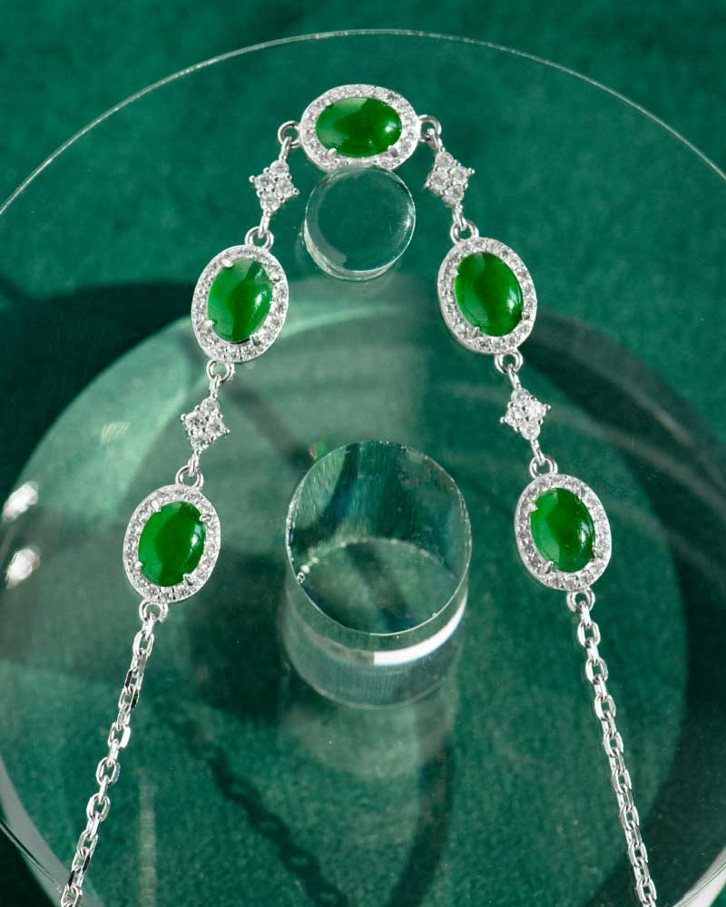LYU_5289 The Vivid Green Jadeite Halo Link Bracelet features domed green cabochons in high-polish sterling silver, arranged symmetrically and displayed on a round glass stand over a green background.