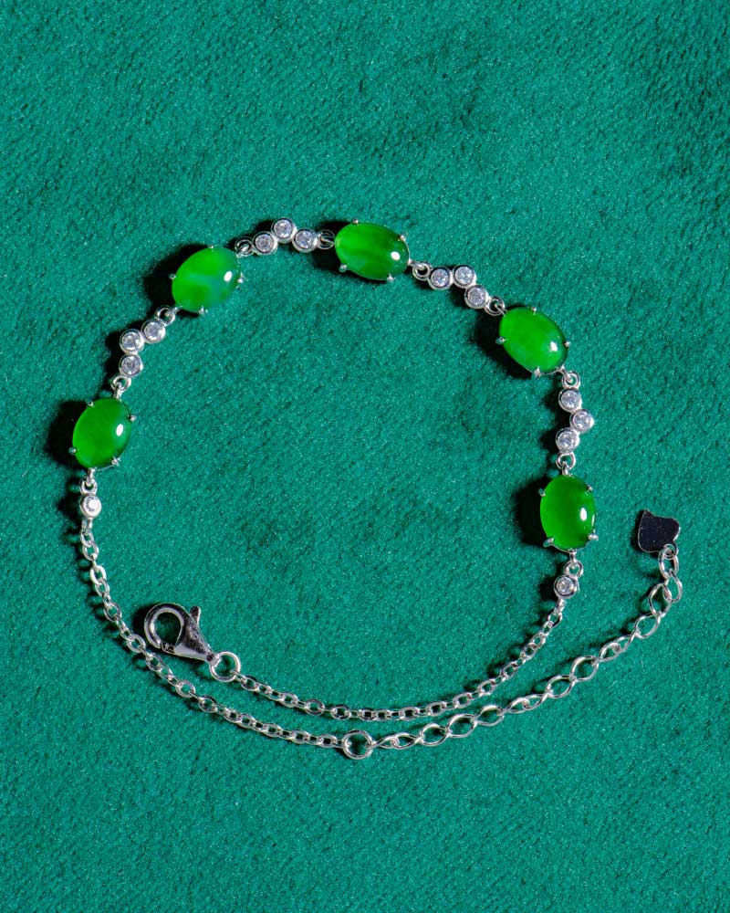 The Vivid Green Jadeite Link Bracelet, featuring five oval cabochons in high-polish sterling silver, is displayed on a textured green fabric background.