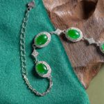 The Vivid Green Jadeite Halo Link Bracelet, featuring oval domed cabochons in high-polish sterling silver, is shown on green fabric and brown textured wood with its adjustable chain visible.