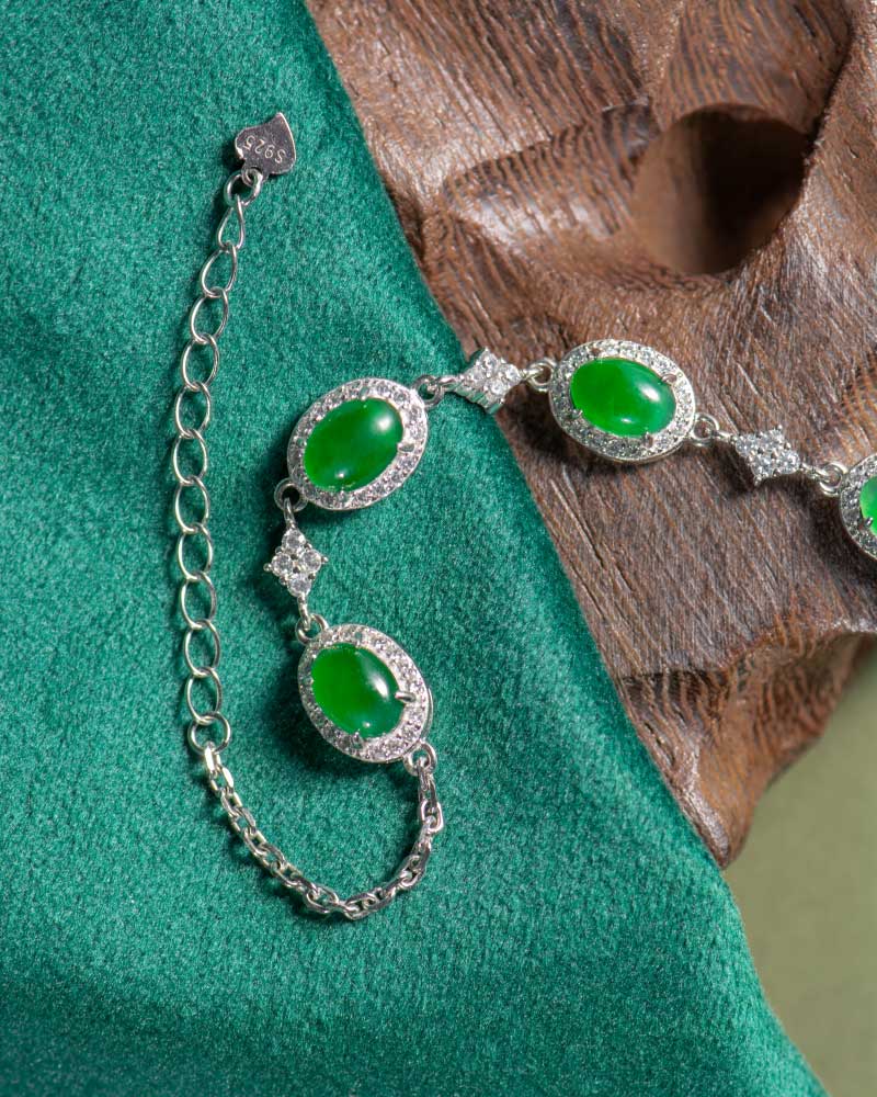 LYU_5305 The Vivid Green Jadeite Halo Link Bracelet, featuring oval domed cabochons in high-polish sterling silver, is shown on green fabric and brown textured wood with its adjustable chain visible.