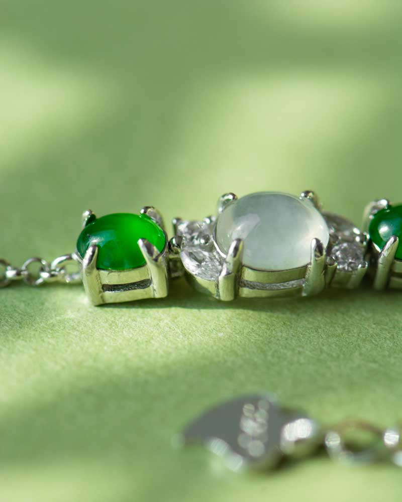 LYU_5324 Close-up of the Green & Greenish White Jadeite Halo Station Bracelet—oval cabochons set in high-polish sterling silver—featuring green and translucent gemstones, on a light green surface with a softly blurred chain and clasp.