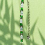 The Green & Greenish White Jadeite Halo Station Bracelet features alternating oval cabochons set in high-polish sterling silver, an adjustable chain, and a delicate heart charm, shown resting on a light green surface with leafy shadows.