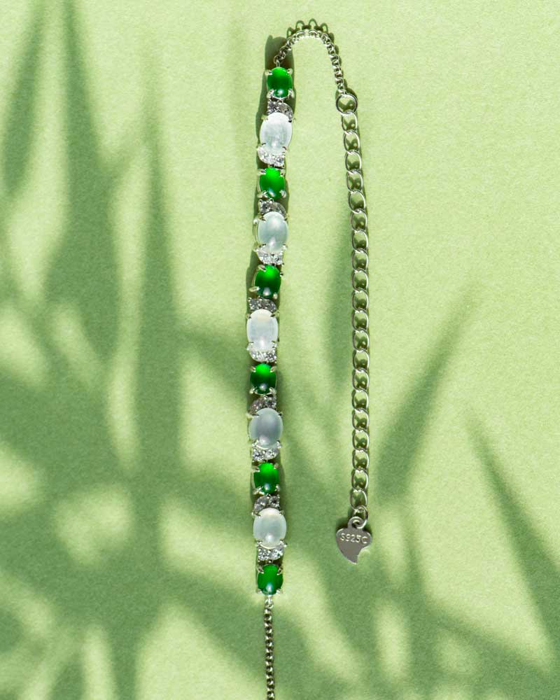LYU_5328 The Green & Greenish White Jadeite Halo Station Bracelet features alternating oval cabochons set in high-polish sterling silver, an adjustable chain, and a delicate heart charm, shown resting on a light green surface with leafy shadows.