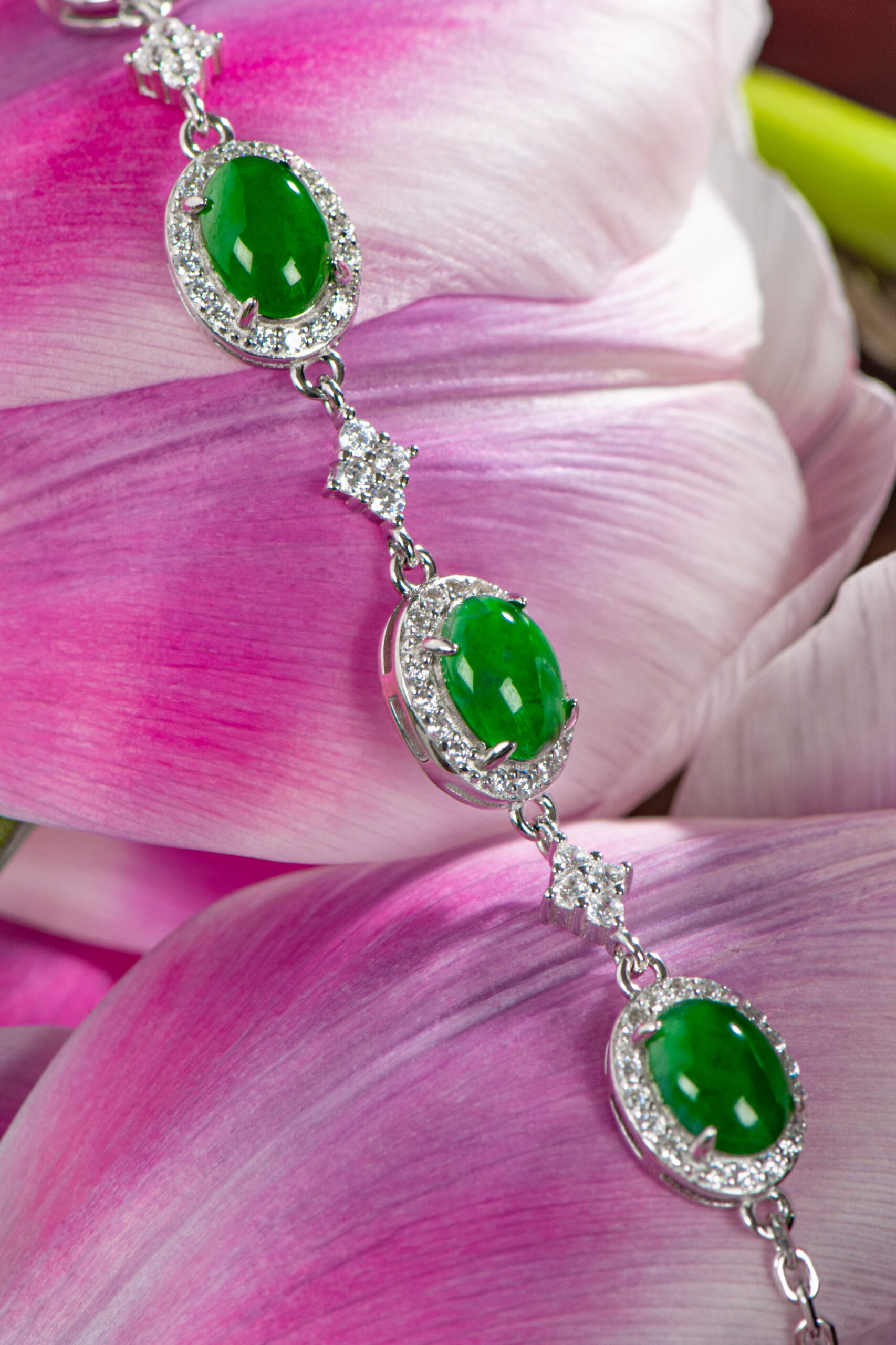 The Vivid Green Jadeite Halo Link Bracelet, with three large oval green gemstones encircled by sparkling stones, is elegantly displayed on pink and white flower petals.