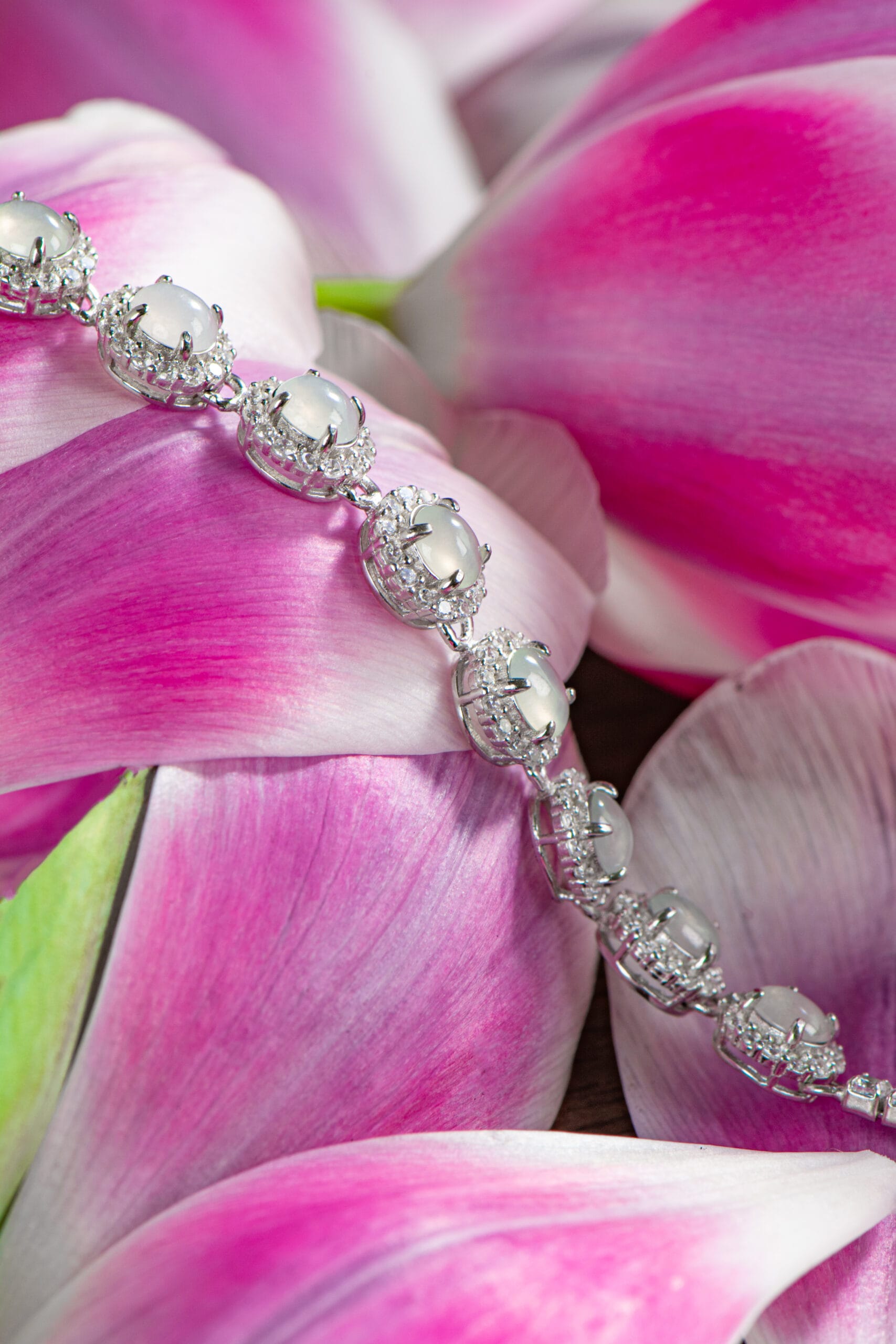 The Icy White Jadeite Tennis Bracelet, featuring oval cabochon gemstones set in 925 sterling silver, is displayed on overlapping pink and white flower petals.