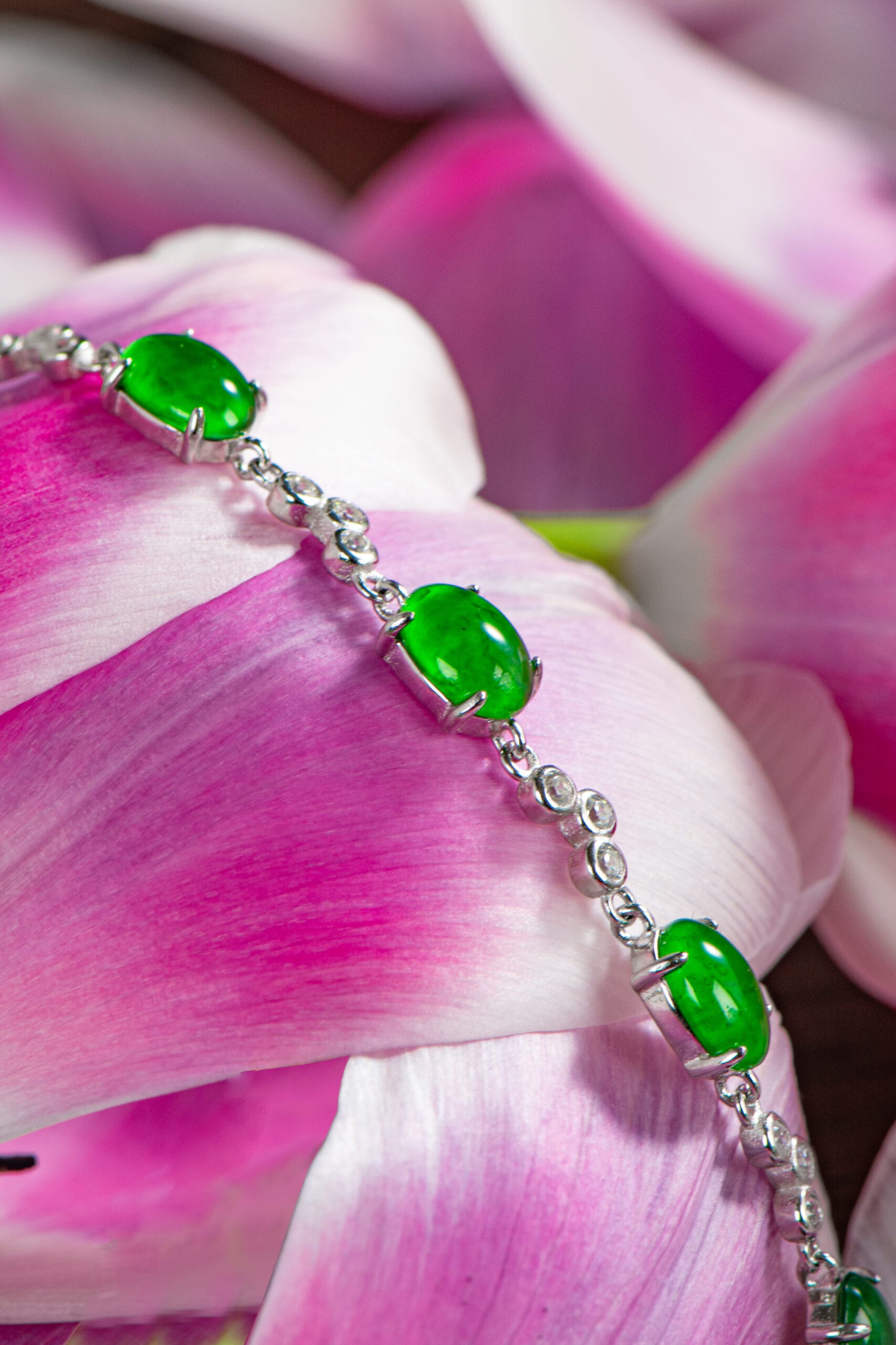 The Vivid Green Jadeite Link Bracelet, featuring oval cabochons set in high-polish sterling silver, rests elegantly across delicate pink and white flower petals.