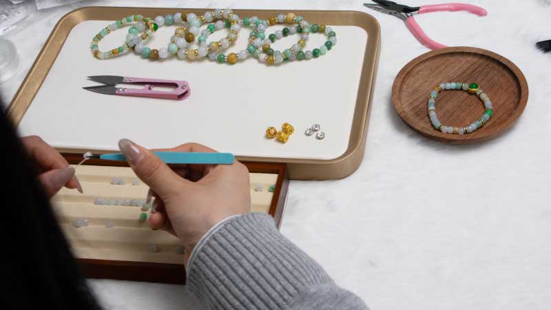 A person uses tweezers to assemble beads for jewelry on a table with finished beaded bracelets, tools, and small containers of beads nearby.